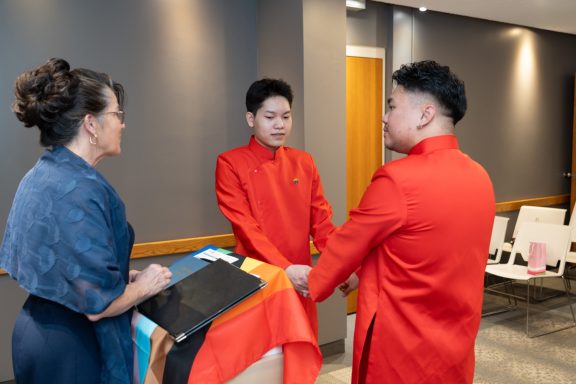 Two men in red attire holding hands, with a woman in blue nearby, in a ceremonial setting.