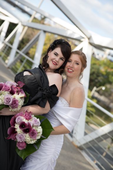 Two brides in elegant dresses hold bouquets of flowers, smiling on a scenic bridge.