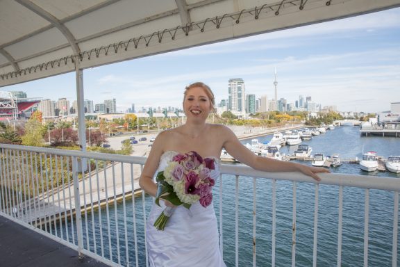 Bride in a strapless white dress holds a bouquet by a marina with city skyline in the background.