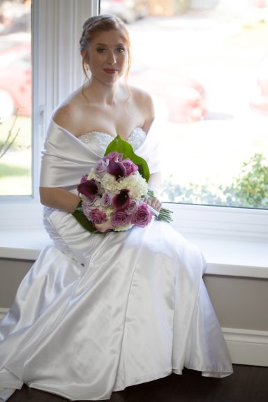 Bride in a white gown holding a bouquet of white and purple flowers, sitting by a window.