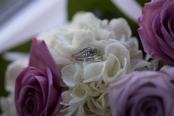 An engagement ring nestled in a bouquet of white and purple roses.