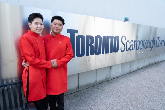 Two individuals in red outfits stand together in front of a Toronto Scarborough sign.